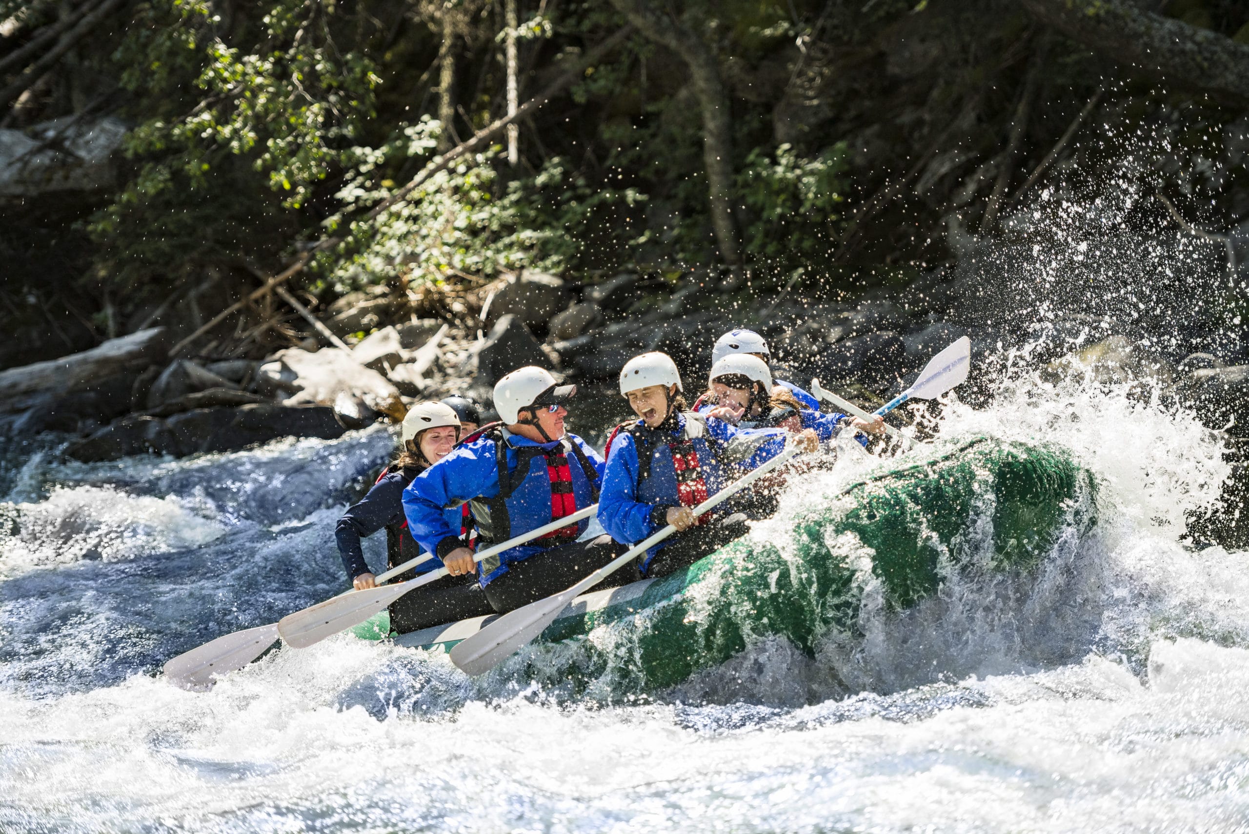 Five people navigate the stunning rapids in their green boat and white hats as the sun shines down on them and the water splashes up happily during their white water rafting trip to Montana with Adventures By Disney.