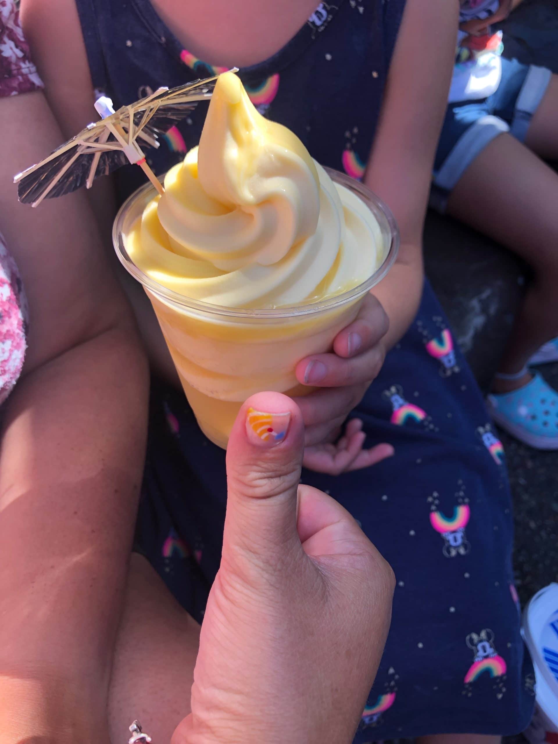 With a colorful-flair-bedecked thumb giving a big thumbs-up in the foreground, the additional hands of someone wearing a blue and pink dress lovingly clasp the invitingly yellow Pineapple Dole Whip that is one of the most beloved staple treats of Disneyland California for visiting families of all shapes, sizes and backgrounds (especially with its cute little umbrella topper.)