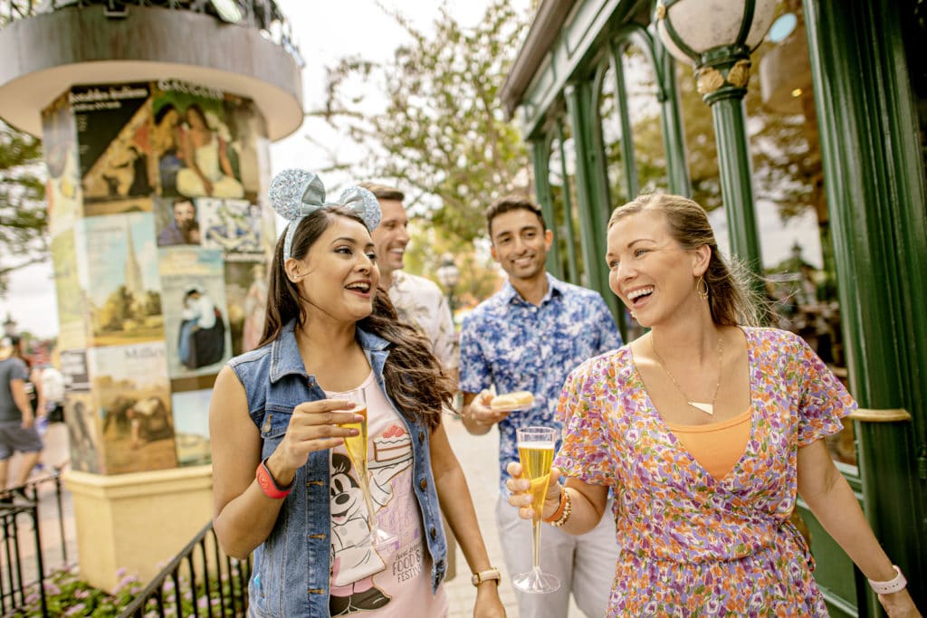 Two women (one with sparkly Mickey ears) and two men walk through Epcot enjoying drinks and snacks and showing how much fun grown-ups can have at Disney World too.