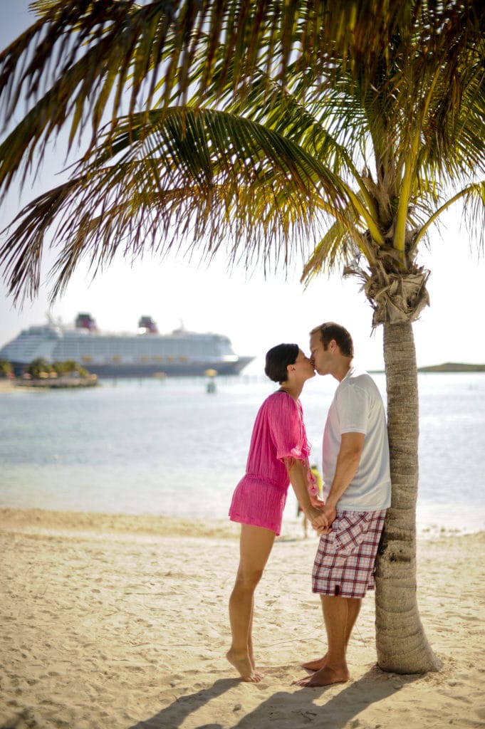 A man and woman kiss in the foreground, pressed adorably against a palm tree, with a Disney cruise ship in the background, as they enjoy the endless romantic possibilities of a Disney honeymoon