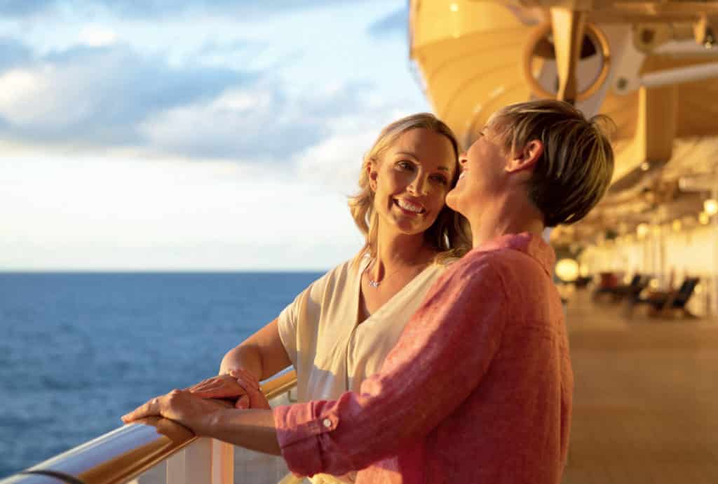 Two completely in love women hold hands and laugh together while enjoying the beautiful sunset on their Disney cruise honeymoon