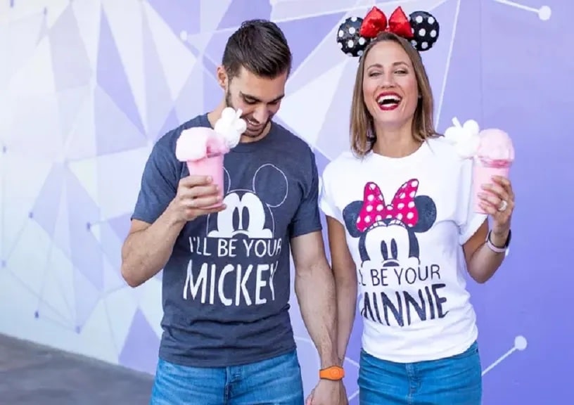 A man wearing an "I'll Be Your Mickey" shirt holds a Disney treat in one hand and his new wife wearing an "I'll Be Your Minnie" shirt in the other hand as they happily laugh and walk around during their honeymoon at Disney World